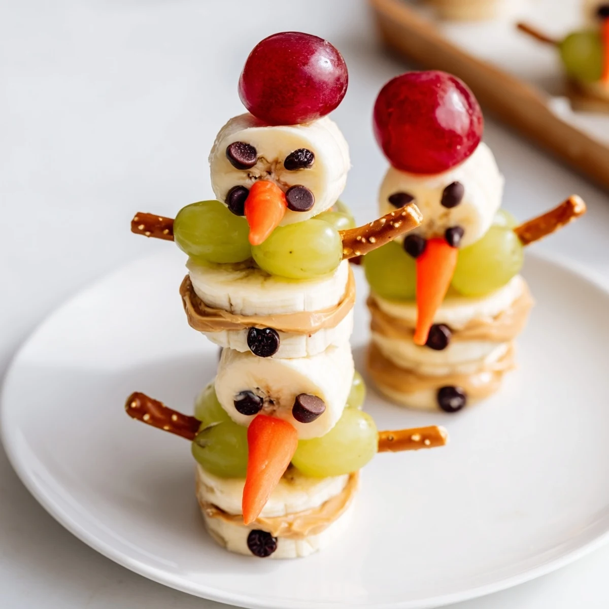 Adorable banana peanut butter snowmen arranged, featuring chocolate chip eyes and festive orange noses for a holiday snack.