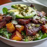 Caramelized butternut squash steak bowl topped with creamy avocado and pumpkin seeds over greens.