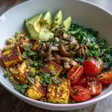 Colorful vegan breakfast bowl features turmeric tofu scramble, savory mushrooms, bright green kale, and couscous topped with creamy avocado and cherry tomatoes. 